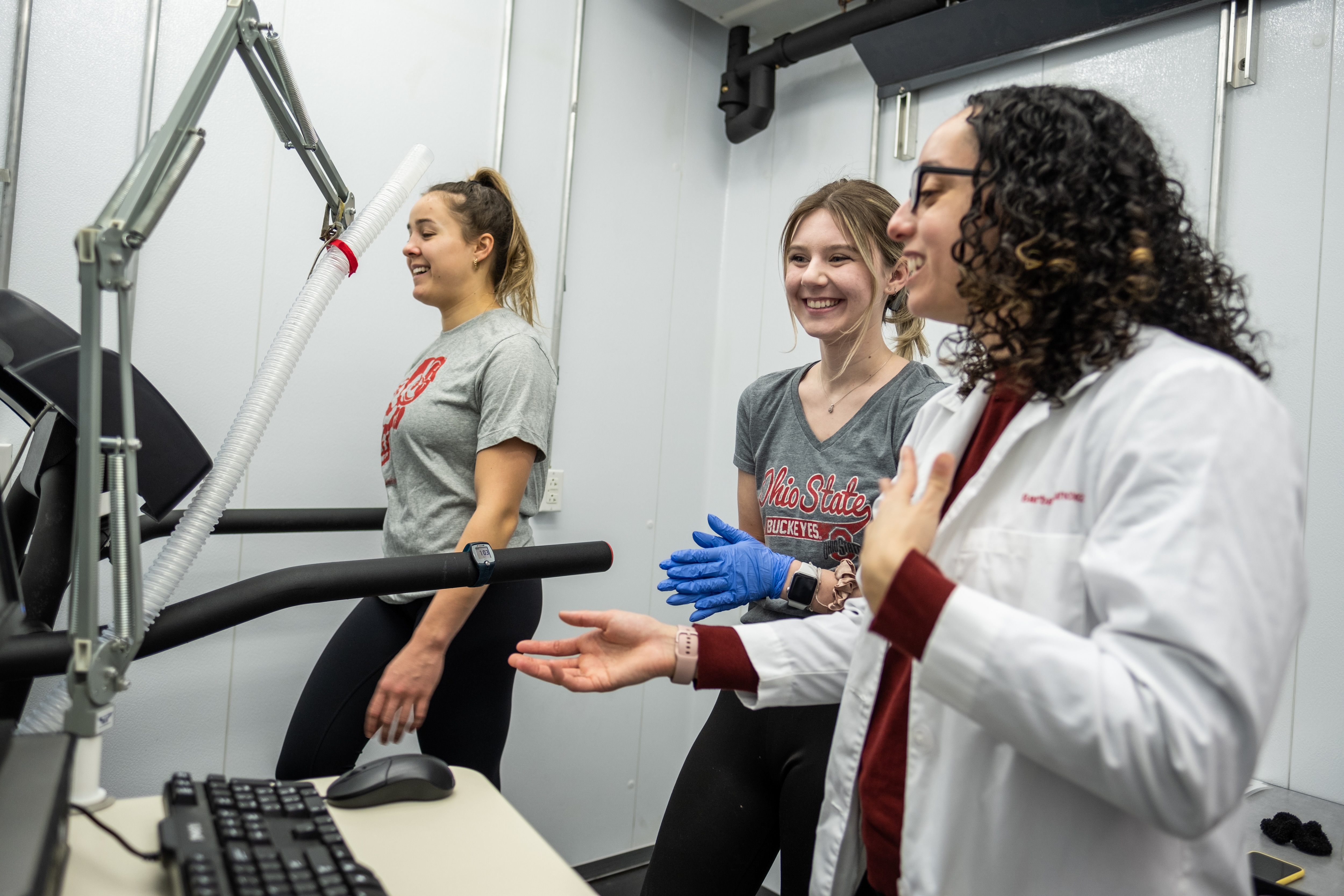 A person wearing an Ohio State Buckeyes T-shirt stands on a treadmill inside a lab while another person in gloves and a third person in a white lab coat observe and discuss. A computer monitor and tubing connected to the treadmill are visible in the foreground.